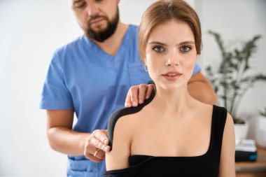 Kinesiyoterapi kliniğindeki bir erkek, mavi üniformalı bir adamın omzuna bant yapıştırır.