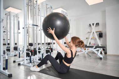 Bir kadın spor salonunda spor yaparken, bir fitball topuyla egzersiz yapar.