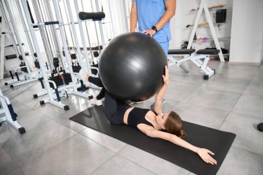 Takım elbiseli bir kadın spor salonunda antrenör gözetiminde bir fitball topuyla çalışıyor.