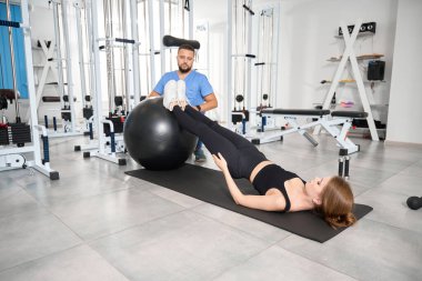 Genç bayan antrenör gözetiminde bir fitball topuyla egzersiz yapıyor.