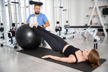 Kinesiyoterapi kliniğindeki hasta, eğitmenin gözetiminde bir fitball topuyla egzersiz yapar.