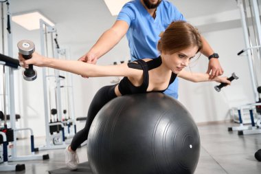 Güzel bir kadın, eğitmenin gözetiminde bir fitball ve dambıllarla egzersiz yapıyor.