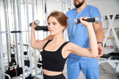 Genç bir kadın, fizyoterapist gözetiminde dambıllarla egzersiz yapıyor. Antrenman kıyafeti giymiş bir kadın.