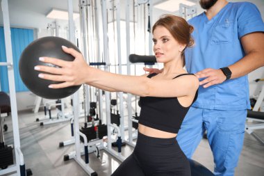 Hasta, bir fizyoterapistin gözetiminde topla egzersiz yapıyor. Antrenman kıyafetli bir kadın.