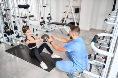 Hasta, mavi üniformalı bir adam olan kineziyologun gözetiminde bir topla egzersiz yapıyor.