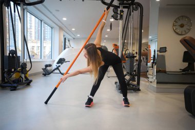 Antrenman kıyafeti giymiş güzel bir kadın fiziksel esneme egzersizleri yapıyor, özel spor sopaları kullanıyor.