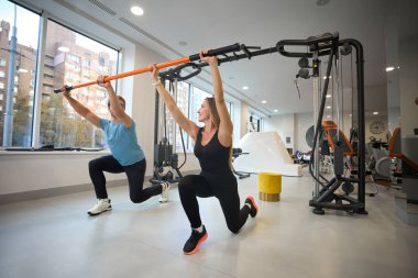 Orta yaşlı bir kadın ve erkek özel bir sopayla ısınıyorlar. Modern bir spor salonunda çalışıyorlar.