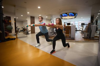 Erkek ve kadın halterlerle egzersiz yapıyorlar. Modern bir spor salonundalar.