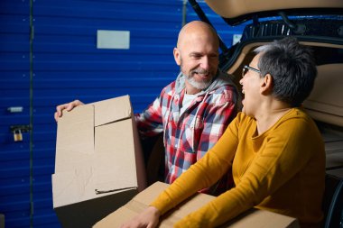 Bir adam ve bir kadın ellerinde bir kutuyla bir arabanın bagajında oturuyorlar.