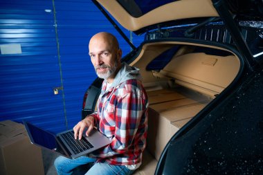 Sakallı adam elinde dizüstü bilgisayarla bagajın kenarında oturuyor..