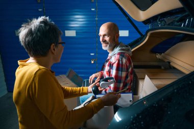 Bir erkek ve bir kadın, bir arabanın bagajının yanında bir adamla iletişim kuruyor.