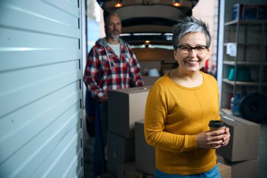 Bir adam ve bir kadın, bir arabanın yanındaki depoda dikiliyorlar.