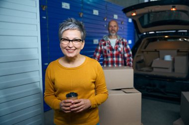 Kadın elinde bir bardak kahveyle kameraya bakarken bir adam elinde karton bir kutu tutuyor.