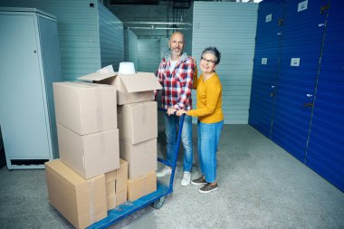 İki yetişkin, içinde katlanmış şeyler olan karton kutuları taşır.