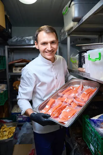 Man stands with a tray of red fish in a restaurant refrigerator, surrounded by shelves of food