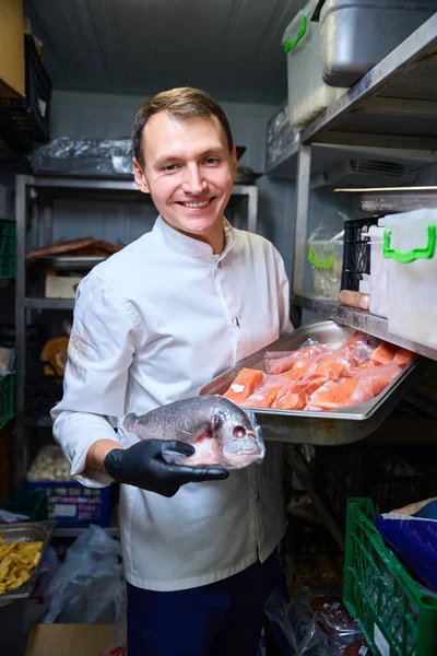 Cook stands with fish in his hands in a restaurant refrigerator, there are shelves of food around him