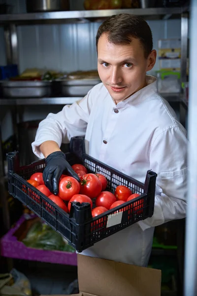 男はレストランの冷蔵庫にトマトの箱を持ってきた その周りに食べ物の棚があります — ストック写真