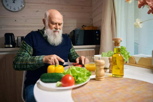Gray-bearded old man at the kitchen table is chopping vegetables for a salad, he is wearing a warm blue vest