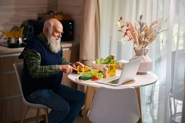 Old man is preparing a salad and chatting online, he is wearing a warm blue vest