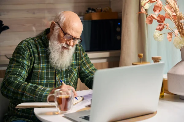 Yaşlı adam mutfak masasında dizüstü bilgisayarda çalışıyor. Masanın üzerinde kurumuş çiçek kompozisyonu var.