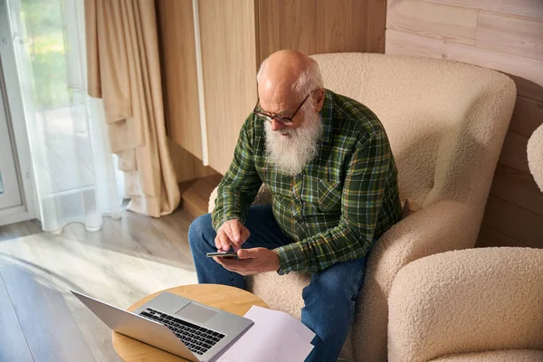 Gri saçlı yaşlı adam oturma odasında belgelerle çalışıyor. Dizüstü bilgisayar ve cep telefonu kullanıyor.