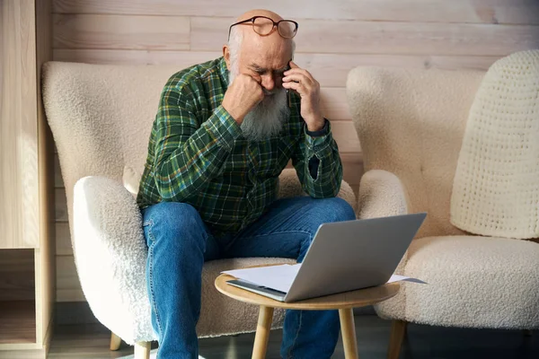Old man is tired, working with documents in the living room, he communicates on his mobile phone