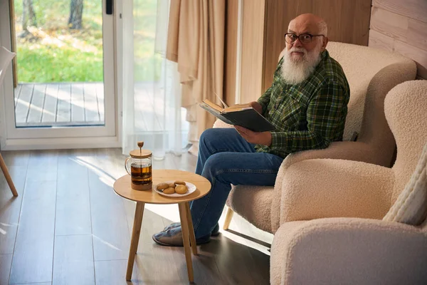 Gray-bearded old man in glasses settled down with a book by the French window, he sits in a comfortable chair