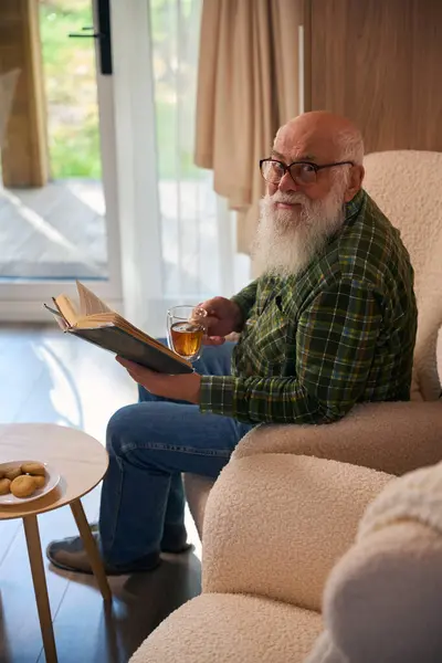 Old man with glasses settled down with a book by the French window, he sits in a comfortable chair