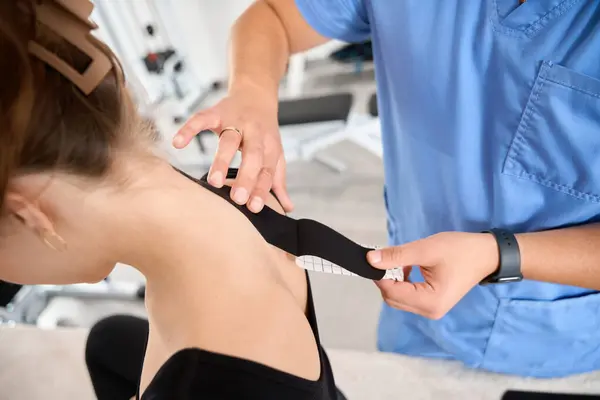 Specialist kinesiologist glues black tape to the back of a patient, a woman in comfortable black clothes