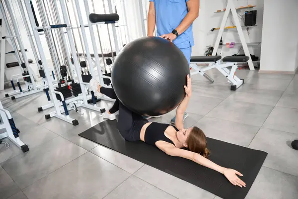 Takım elbiseli bir kadın spor salonunda antrenör gözetiminde bir fitball topuyla çalışıyor.
