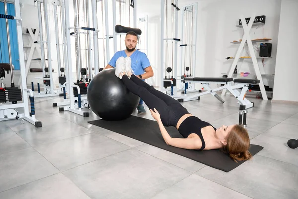 Genç bayan antrenör gözetiminde bir fitball topuyla egzersiz yapıyor.