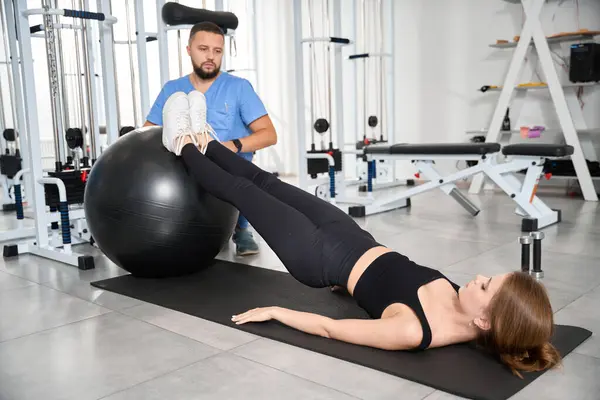 Kinesiyoterapi kliniğindeki hasta, eğitmenin gözetiminde bir fitball topuyla egzersiz yapar.