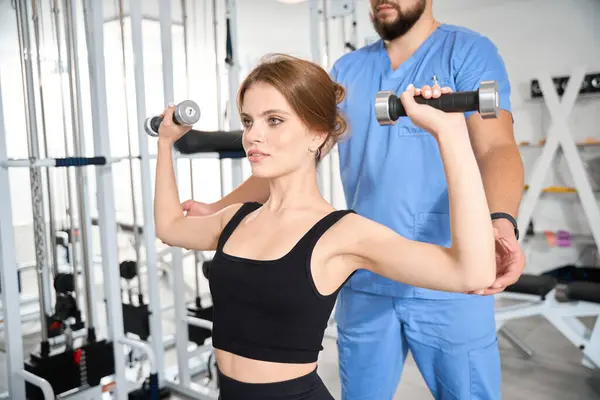 Genç bir kadın, fizyoterapist gözetiminde dambıllarla egzersiz yapıyor. Antrenman kıyafeti giymiş bir kadın.