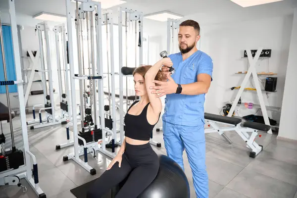 Güzel bir kadın, fizyoterapistin gözetiminde dambıllarla egzersiz yapar. Kadın, formda bir topun üzerine oturur.