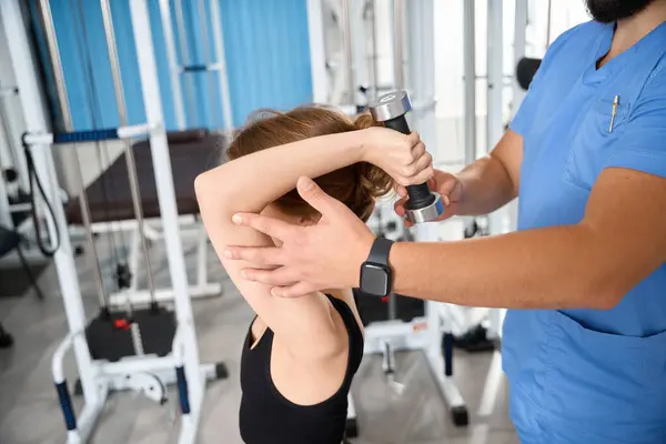 Kadın, mavi üniformalı bir eğitmenin gözetiminde dambıllarla egzersiz yapıyor.