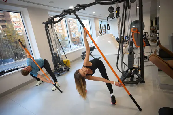 Man and woman in training clothes perform physical exercises in the gym, sports sticks are used