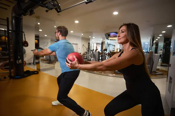 Kadın ve erkek bir topla egzersiz yapıyorlar, modern bir spor salonundalar.