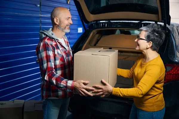 Kel sakallı adam gülümseyen bir kadının elinden kutuyu alıyor.