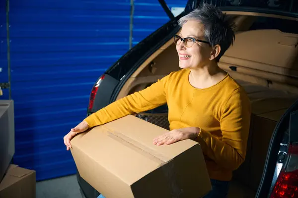 Woman with cardboard box on her lap sitting on car trunk