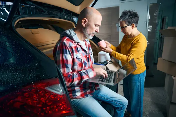 Bir kadın kupaya bakarken elinde dizüstü bilgisayar tutan kel bir adam.