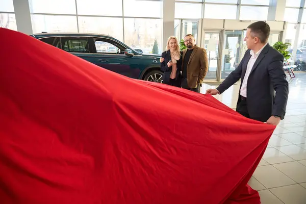 Two people watch a car salesman remove a gift cover from a new car