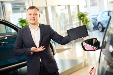 Otomobil satıcısı elinde tabletle kameranın önünde duruyor.