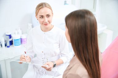 Kadın doktor hastanede oturuyor, hastayı dinliyor ve not alıyor.