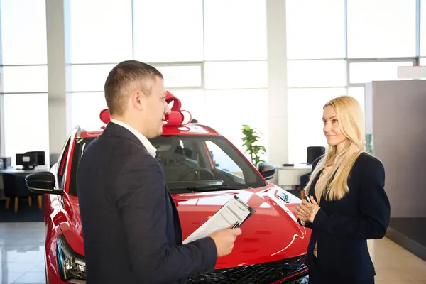 Car salesman talks to a young woman against the background of a red car that was given to her