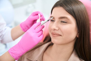 Pembe eldivenli kadın kozmetik uzmanı elinde şırınga tutuyor ve klinikteki bayana gençleştirme iğnesi yapıyor.