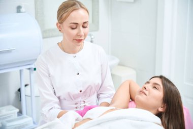 Güzellik salonunda kanepede uzanmış kadın müşteri, kozmetik uzmanı yanında oturuyor ve koluna iğne yapıyor.