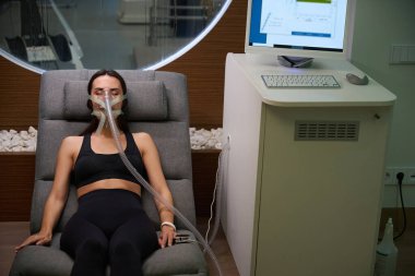 Spor elbiseli bir kadın maske takmış rahat koltukta uzanıyor ve hipoksik terapi görüyor.