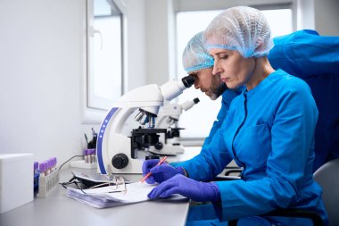 Mavi üniformalı laboratuvar asistanları modern bir laboratuvarda çalışıyorlar. Güçlü bir mikroskop kullanılıyor.