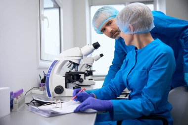 Mavi üniformalı meslektaşlar modern bir laboratuvarda çalışır, güçlü bir mikroskop kullanılır.