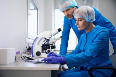 Mavi üniformalı insanlar modern bir laboratuvarda çalışıyorlar. Güçlü bir mikroskop kullanılıyor.